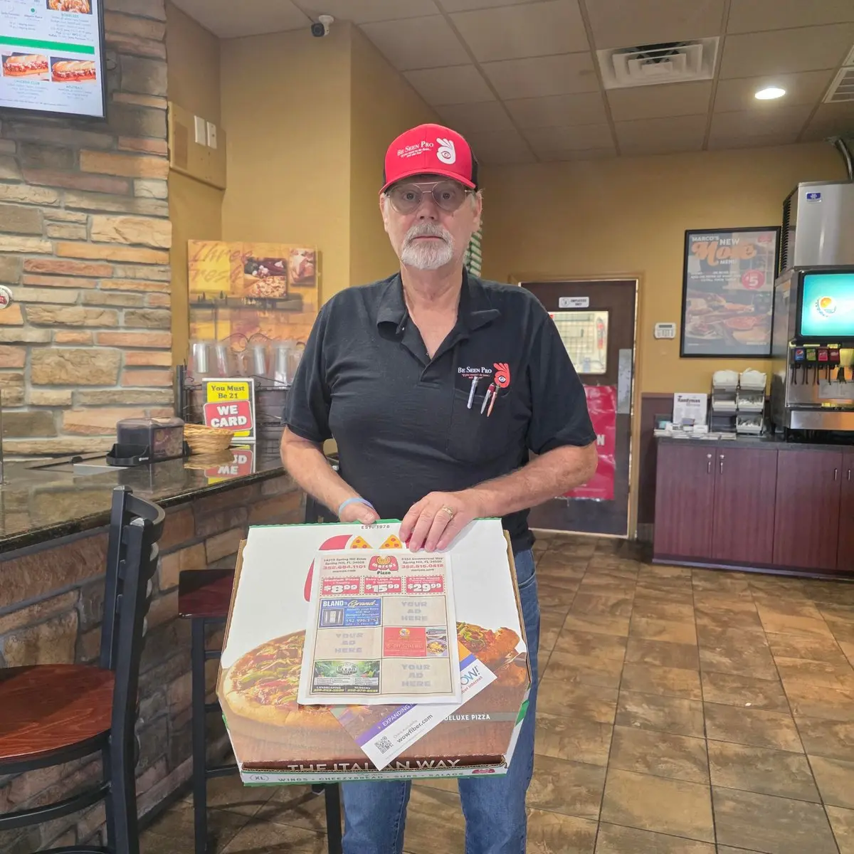 Be Seen Pro representative holding a pizza box topped with a local advertising flyer showing business ads and Your Ad Here spots
