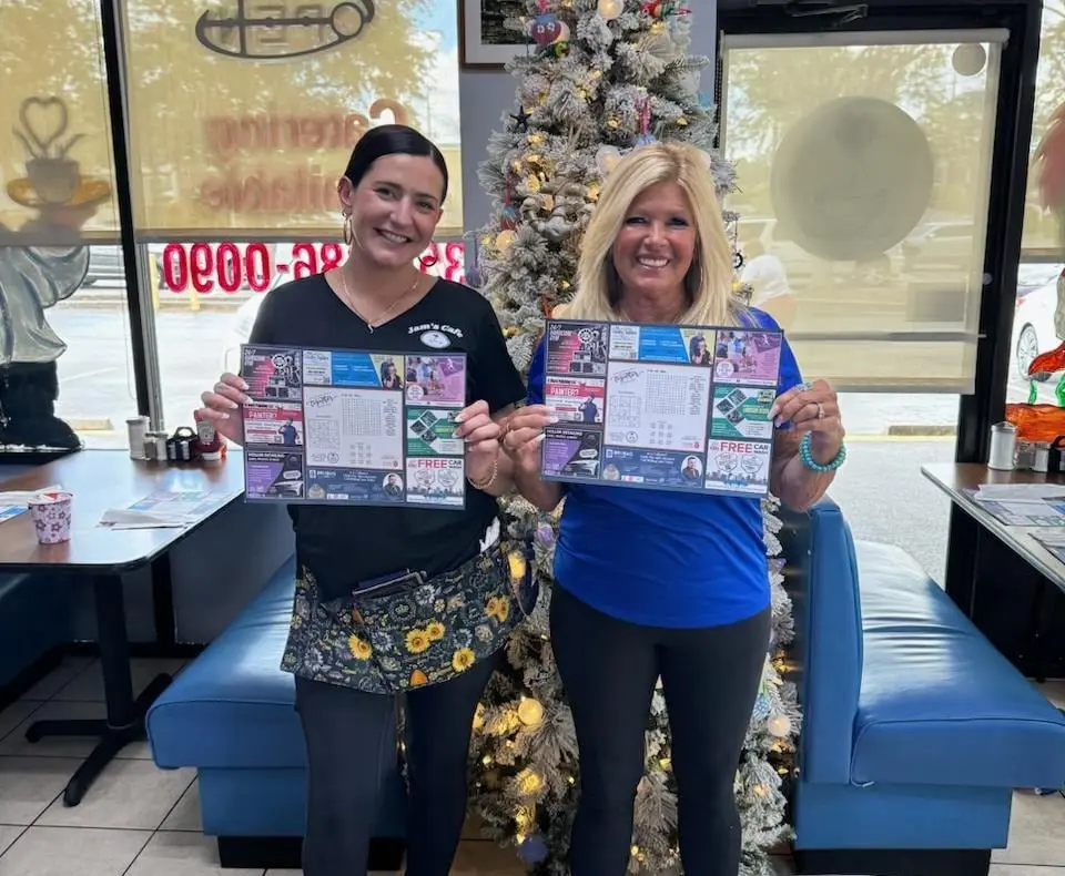 Two smiling Jam's Cafe servers holding Be Seen Pro printed menus in front of a blue booth and decorated Christmas tree